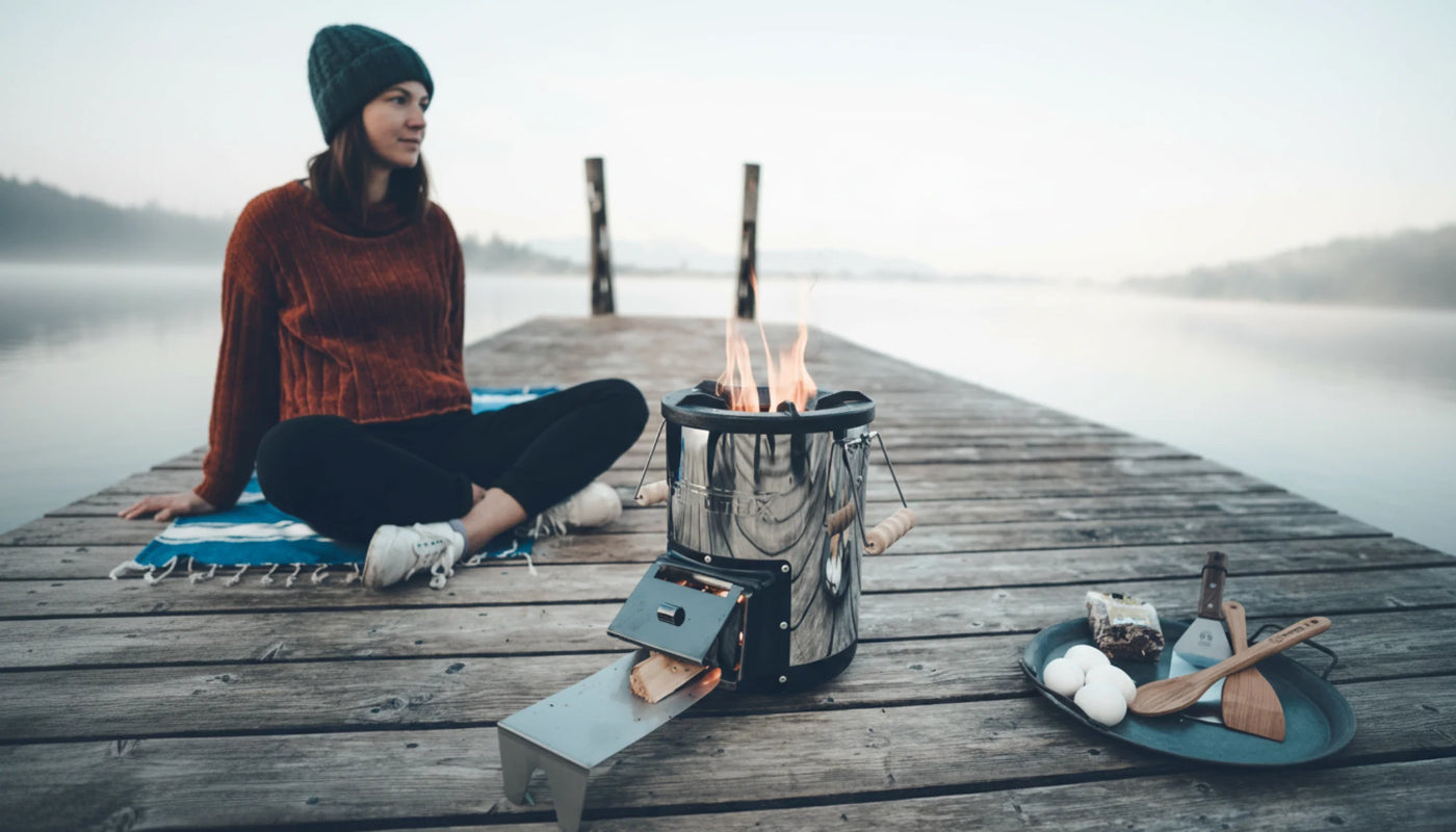 Eine junge Frau sitzt auf einem Steg am See. Im Vordergrund ist der Petromax Raketenofen zu sehen.