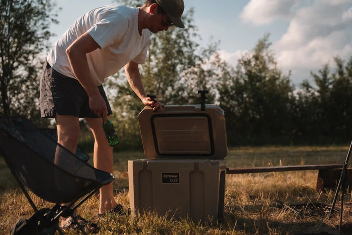Ein Mann legt eine Getränkeflasche in eine geöffnete Petromax Kühlbox auf einem Campingplatz